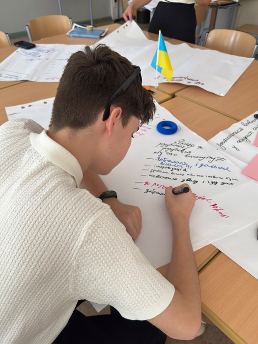 A student writing on a flip-chart sheet in Ukrainian during a resilience exercise, with a small Ukrainian flag on the table.